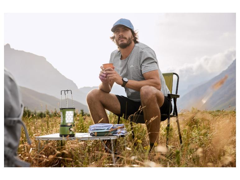 Mann in Kappe und T-Shirt sitzt auf Campingstuhl mit Tasse, daneben Laterne und Tisch.