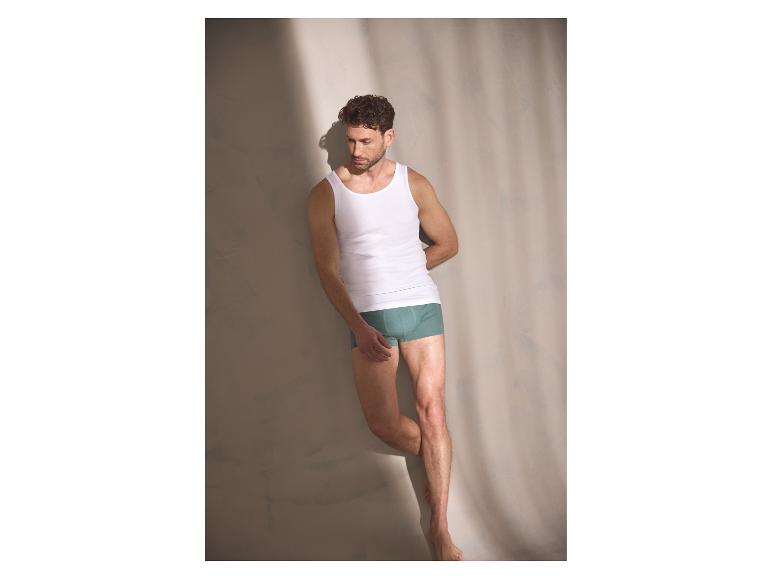 Ein Mann lehnt an einer Wand und trägt Herren-Boxershorts und ein Unterhemd.