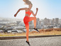 Eine Frau in einem orangen Sport-BH und Leggings springt auf einem Bürgersteig mit Blick auf die Stadt.