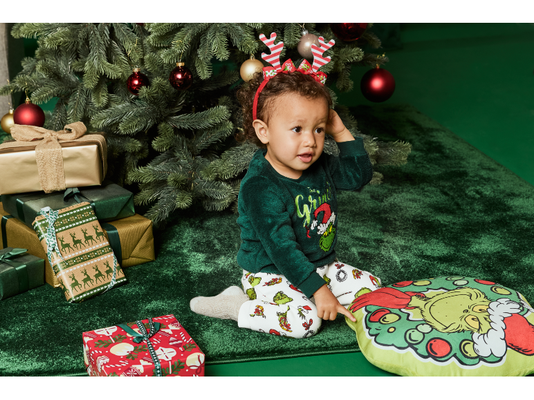 Ein Kleinkind im festlichen Schlafanzug sitzt neben einem Weihnachtsbaum mit Geschenken und einem Grinch-Kissen.