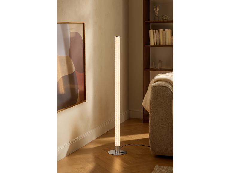 Zylindrische Stehlampe mit Chromfuß beleuchtet ein modernes Wohnzimmer mit Bücherregal und Sofa.