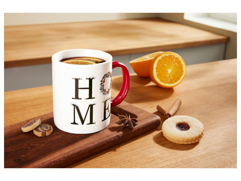 Weiße Tasse mit Feiertagsmotiv, Heißgetränk, Orangen und Süßigkeiten auf einem Holztisch.