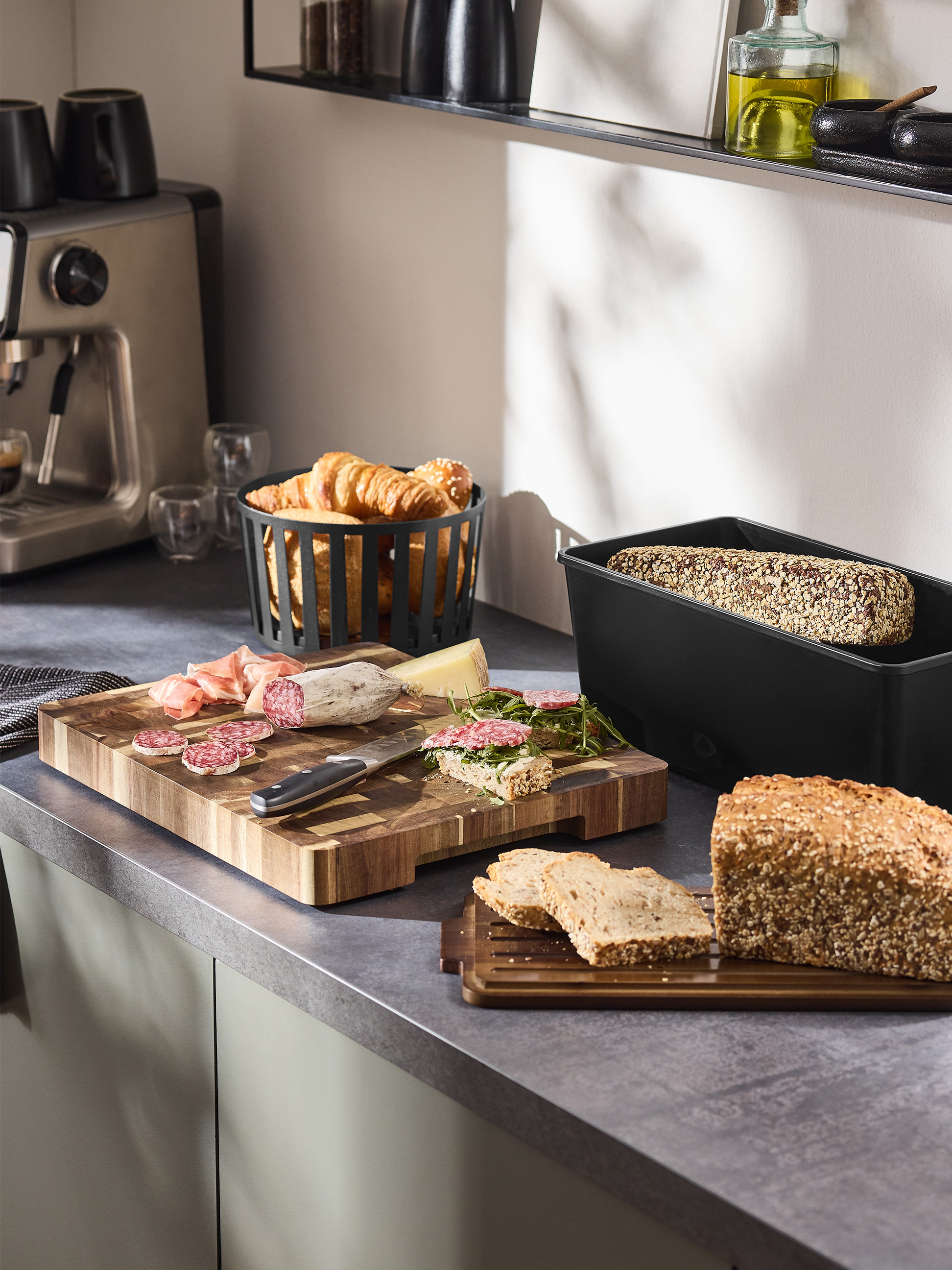 Frühstück mit Kaffeemaschine, Brot, Aufschnitt und Croissants auf einer Küchenarbeitsplatte.