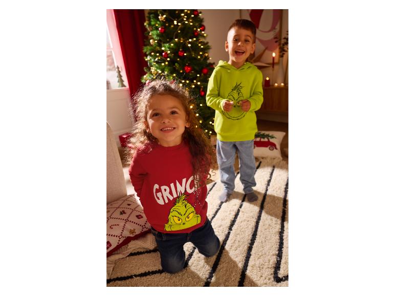 Zwei lächelnde Kinder tragen Grinch-Kapuzenpullover neben einem Weihnachtsbaum.