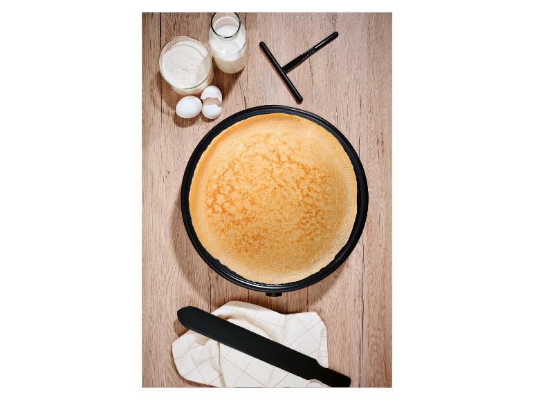 Draufsicht auf ein Crêpes-Eisen mit einem goldenen Crêpe, umgeben von Mehl, Milch und Eiern.