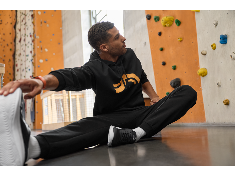 Mann in schwarzem Hoodie und Jogginghose dehnt sich auf dem Boden vor einer Kletterwand.