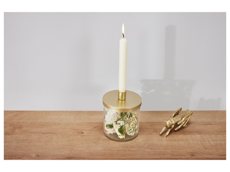 Eine brennende Kerze in einem Glasgefäß mit getrockneten Blumen und eine goldene Heuschrecke auf einem Holztisch.