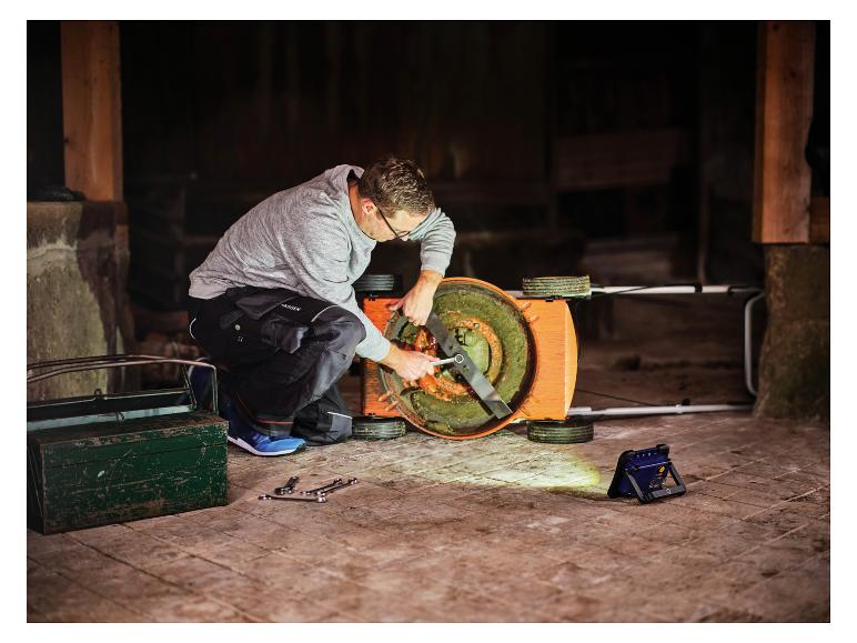 Mann repariert Rasenmäher mit LIVARNO® Arbeitsleuchte »The Portable« in dunklem Schuppen.