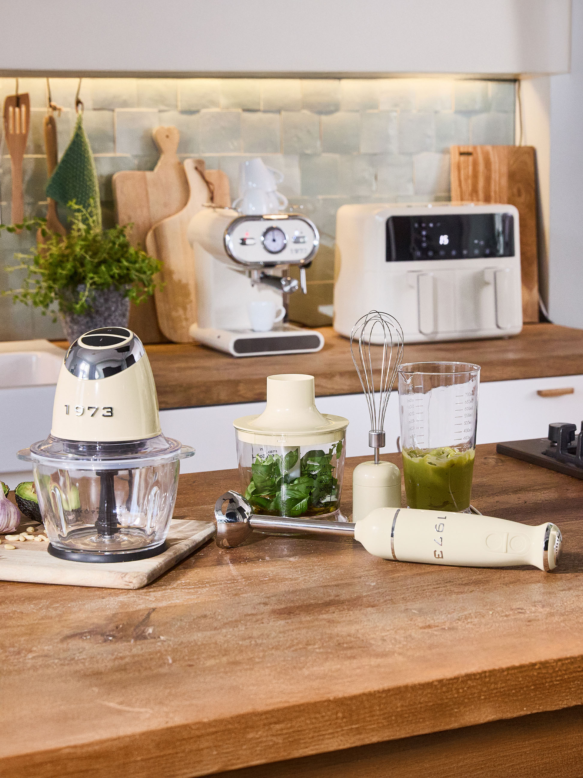 Cremefarbene Küchengeräte, darunter ein Zerkleinerer, Stabmixer und Espressomaschine, auf einer Holztheke.