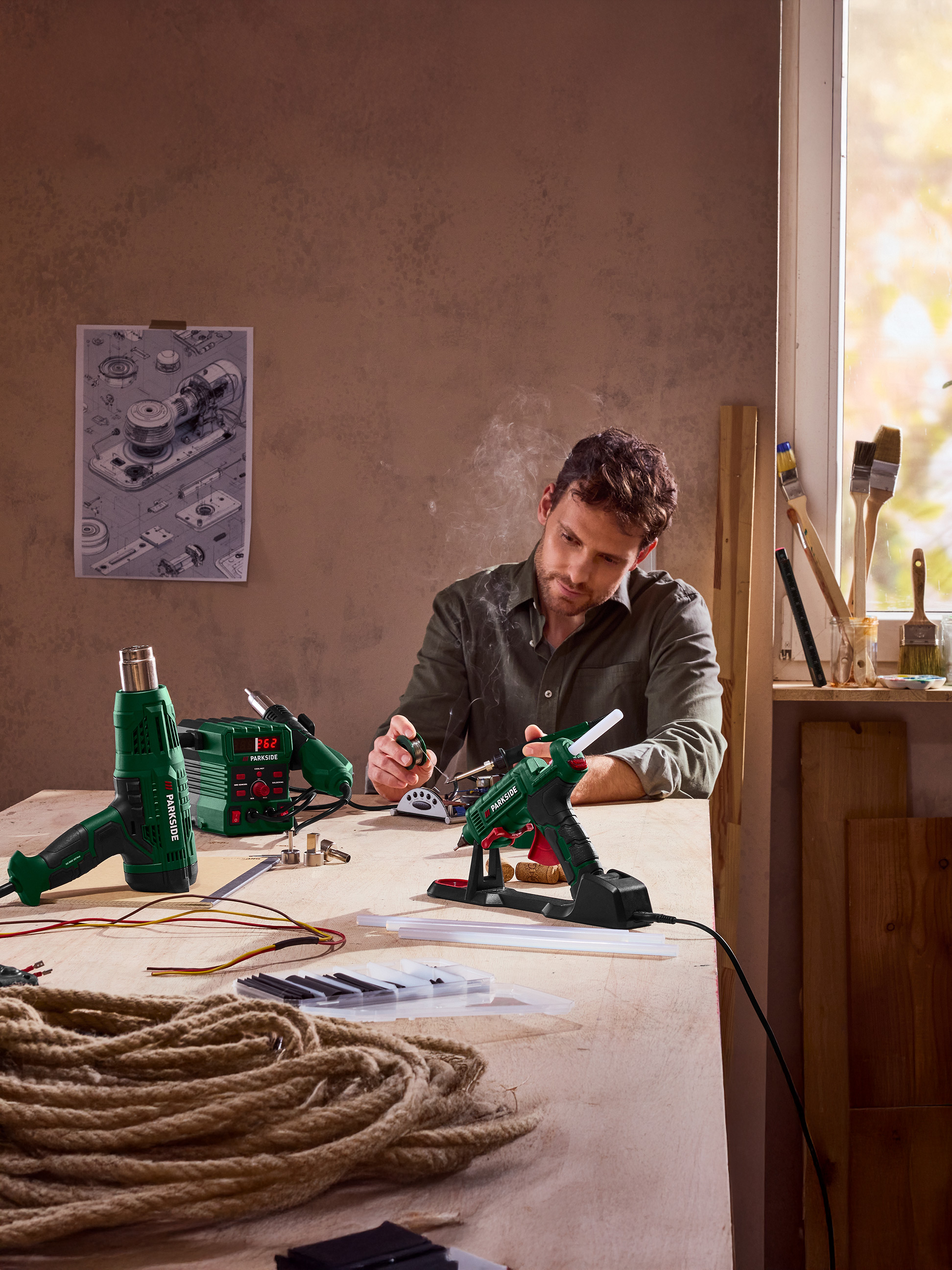 Mann benutzt eine Heißklebepistole, mit Heißluftpistole und Lötkolben auf dem Tisch.