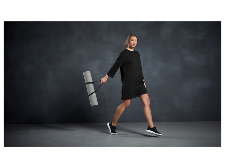 Eine Frau in einem schwarzen Kleid und Sneakern trägt eine Yogamatte.
