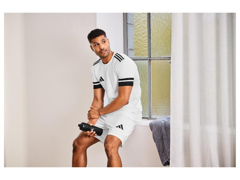 Mann in weißer Adidas Sportkleidung, Shorts und T-Shirt, hält eine Wasserflasche.