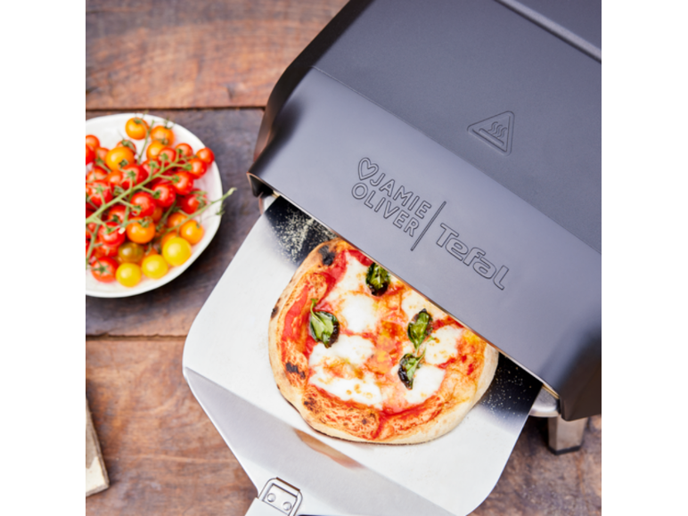 Pizzaofen mit frischer Pizza auf einem Pizzaschieber, daneben Kirschtomaten.
