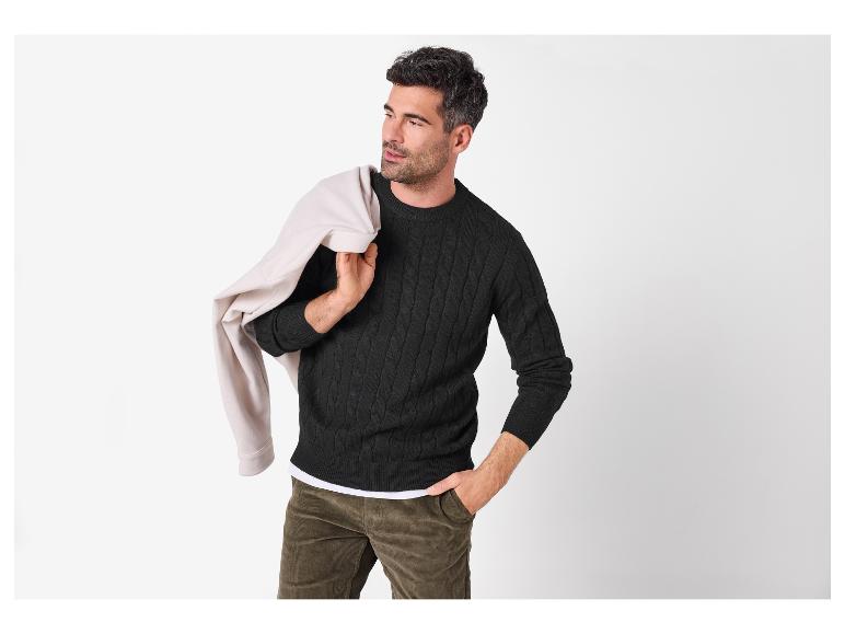 Mann in schwarzem Zopfstrickpullover, Cordhose und beiger Jacke über der Schulter.