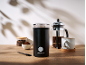 Schwarze elektrische Kaffeemühle, Kaffeebohnen, gemahlener Kaffee und eine French Press auf Holztisch.