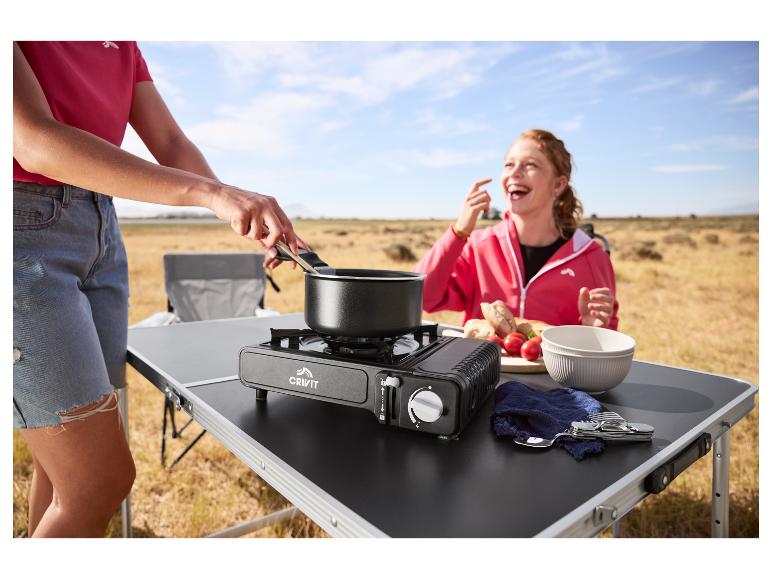 Frau kocht auf einem tragbaren Gaskocher, während eine andere Frau im Hintergrund lacht.