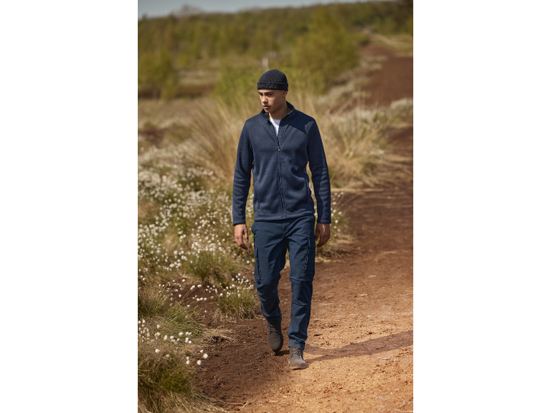 Mann in blauer Fleecejacke, Cargohose und Mütze geht auf einem Feldweg.