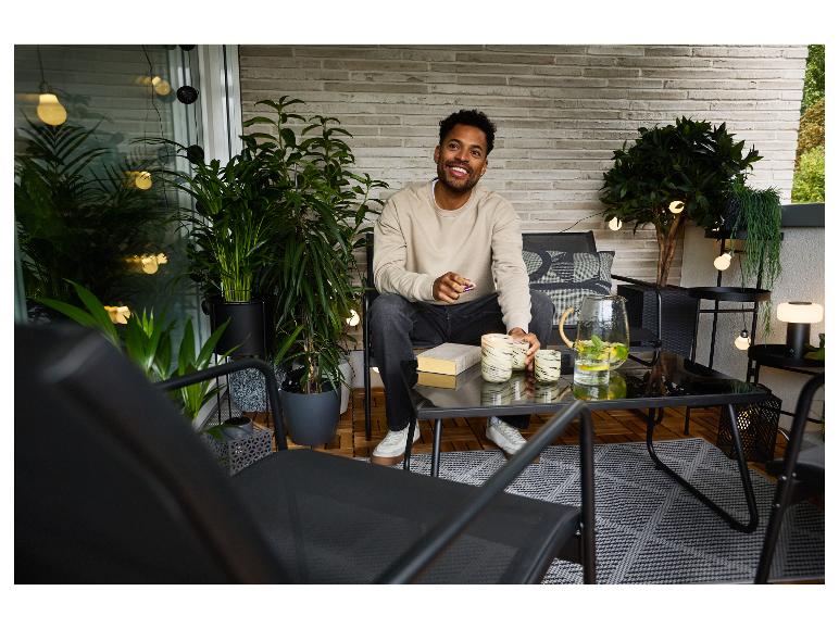 Mann auf Balkon mit Gartenmöbeln, Pflanzen und Lichterketten.