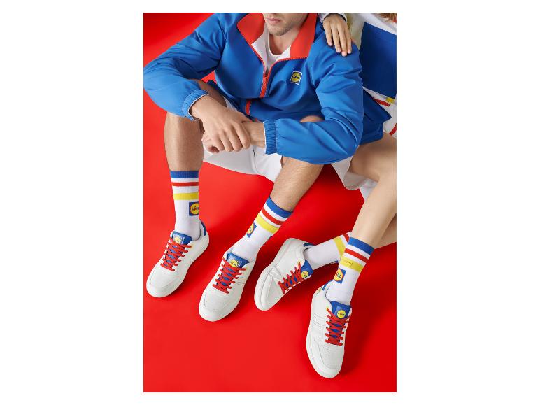 Zwei Personen tragen weiße Sneaker, gestreifte Socken und blaue Jacken vor rotem Hintergrund.