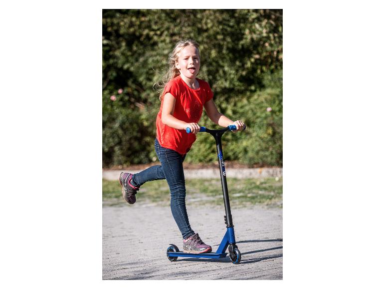 Blondes Mädchen in rotem T-Shirt und blauen Jeans, fährt Roller auf einem Weg.