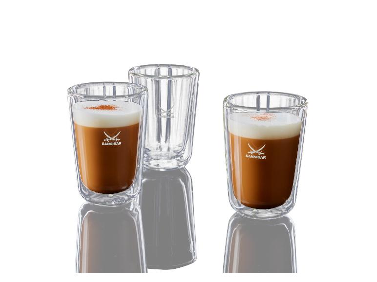 Drei Sansibar-Gläser, zwei gefüllt mit Milchkaffee und eines leer, auf einer reflektierenden Oberfläche.