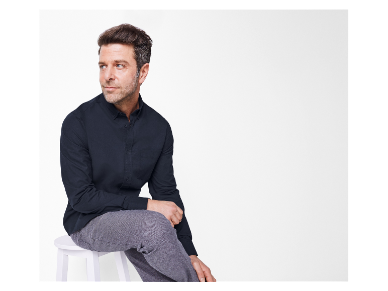 Ein Mann in einem dunkelblauen Hemd und grauer Hose sitzt auf einem weißen Hocker.