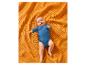 Ein süßes Baby in einem blauen Body liegt auf einer orangefarbenen strukturierten Decke.