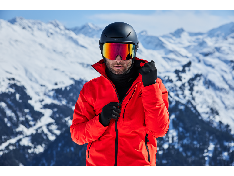 Mann mit Skihelm, Skibrille und roter Jacke in verschneiten Bergen.