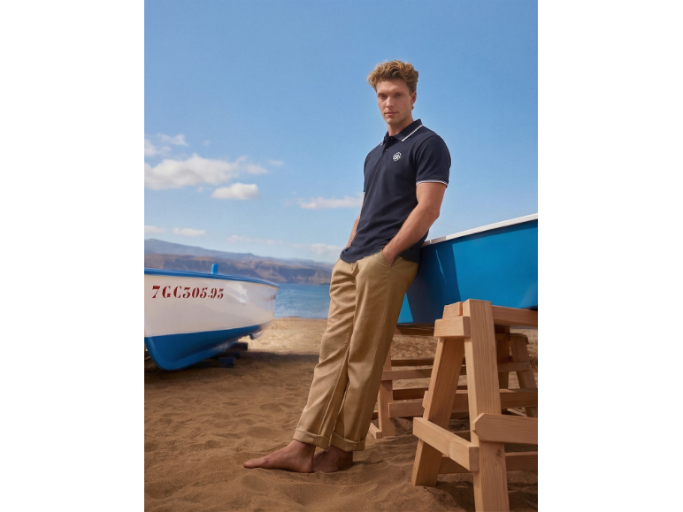 Mann in marineblauem Poloshirt und beiger Hose am Strand mit Booten.