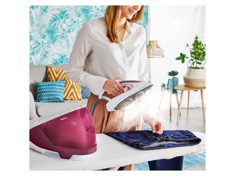 Frau bügelt Jeans mit Dampfbügeleisen, tropische Tapete und Sofa im Hintergrund.