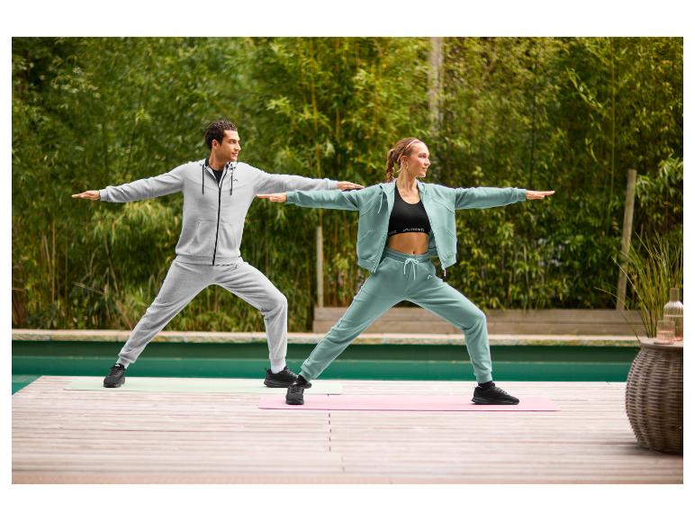 Zwei Personen in Trainingsanzügen und Sportschuhen machen Yoga im Freien auf einer Holzterrasse.