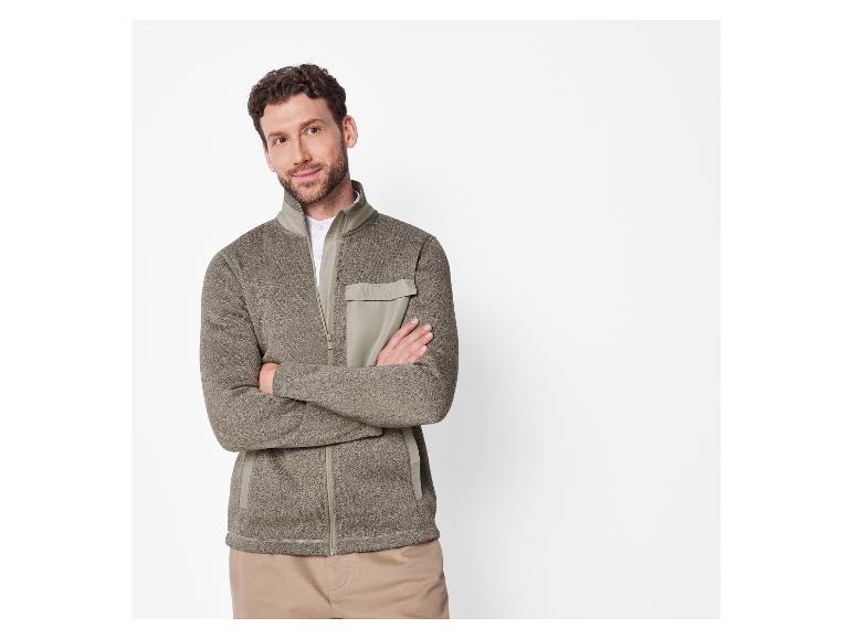 Mann in einer graubraunen Strickjacke mit Reißverschluss und beiger Hose.