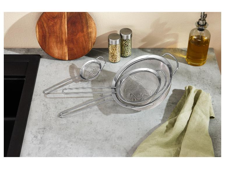 Set aus drei Edelstahlsieben in verschiedenen Größen auf einer Küchenarbeitsplatte mit Schneidebrett und Gewürzen.