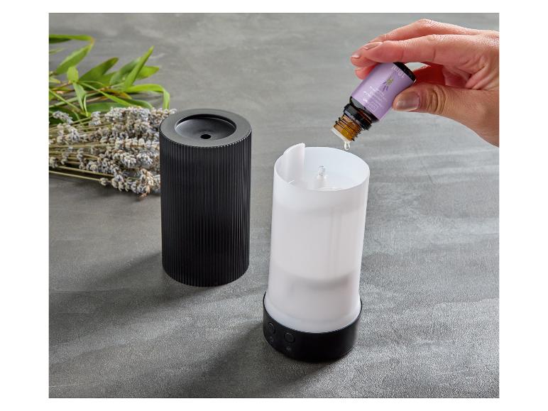 Eine Hand gibt Lavendel-Duftöl in einen Aroma-Diffusor, daneben getrockneter Lavendel und grüne Blätter.