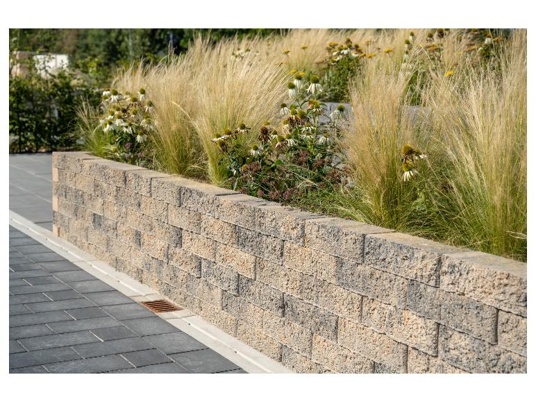 Stützmauer aus Betonblöcken mit Ziergräsern und weißen Blüten.