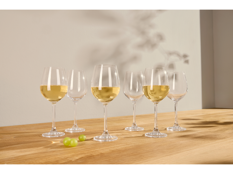 Sechs Weingläser, drei davon mit Weißwein gefüllt, auf einem Holztisch mit Trauben.