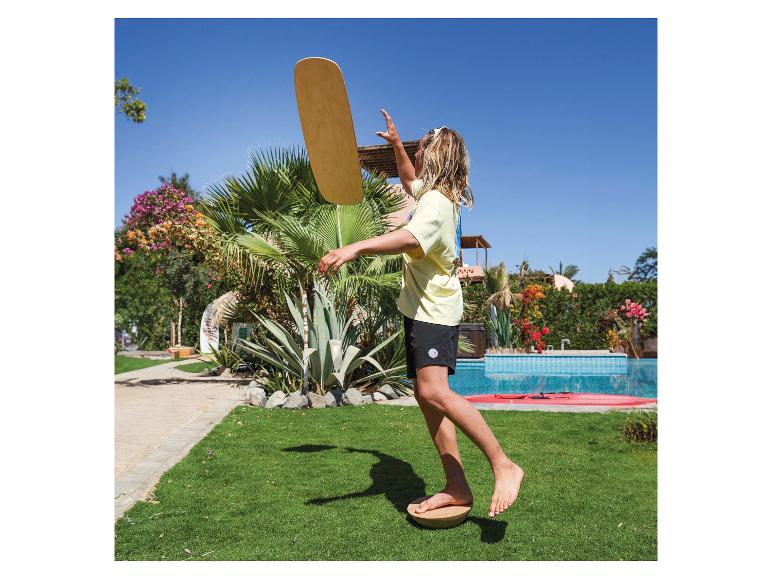 Jugendlicher balanciert auf einem Brett im Freien, neben Pool und Palmen.