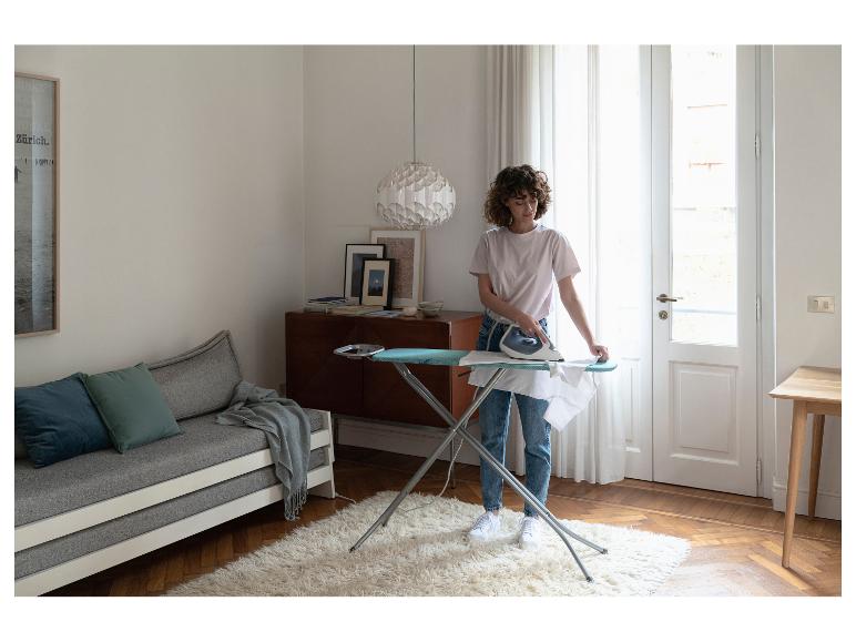 Frau bügelt weißes Hemd auf Bügelbrett im Wohnzimmer mit Sofa und Teppich.