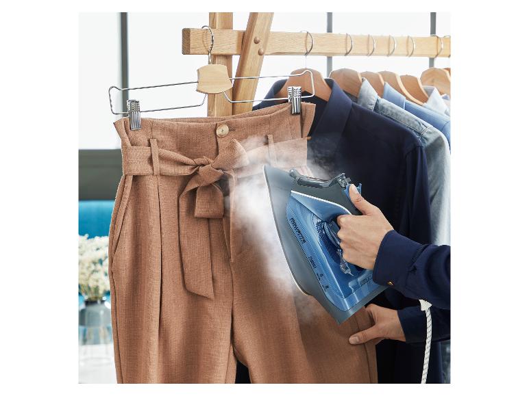 Eine Person dämpft eine braune Hose mit einem blauen Rowenta-Bügeleisen, während Kleidung an einem Ständer hängt.