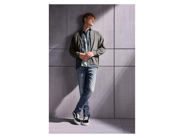 Junger Mann in Jeans, T-Shirt und Bomberjacke lehnt an einer Wand.