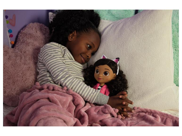 Ein junges Mädchen im gestreiften Pyjama kuschelt eine Puppe mit Katzenohren unter einer rosa Decke.