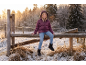 Lächelndes Mädchen in violetter Daunenjacke und Jeans sitzt auf einem Holzzaun in verschneiter Landschaft.