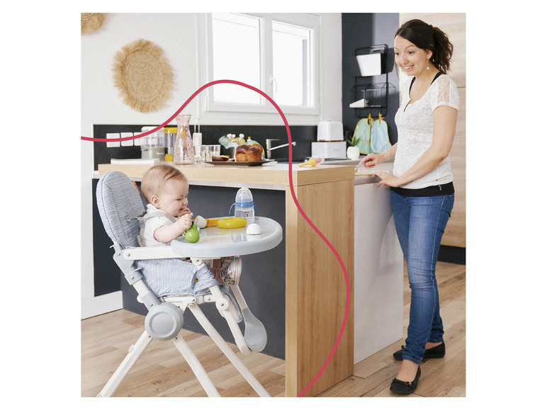 Ein Baby sitzt in einem blauen Hochstuhl und isst, während eine Frau in der Küche steht.