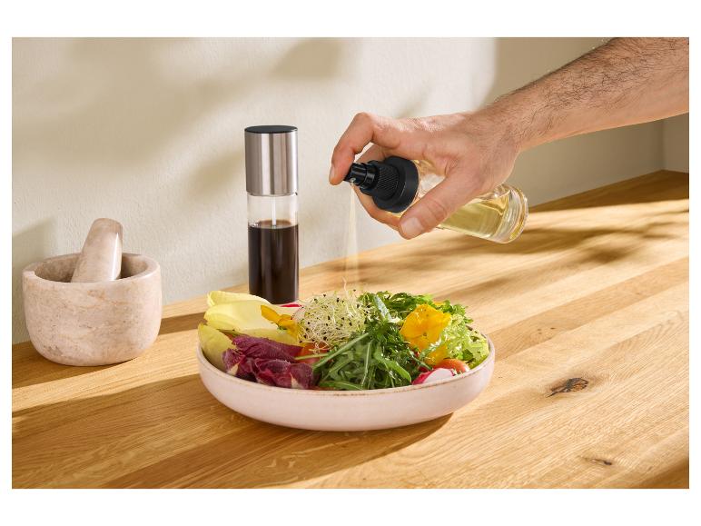 Eine Hand sprüht Öl auf eine Salatschüssel, mit Mörser und Stößel und einer Essigflasche im Hintergrund.
