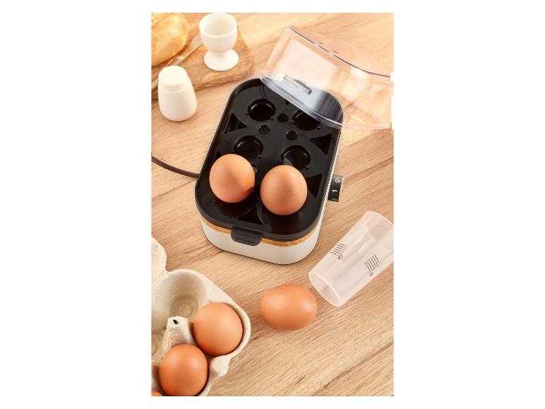 Eierkocher mit Eiern, Messbecher, Salzstreuer, Eierbecher und Brot auf einem Holztisch.