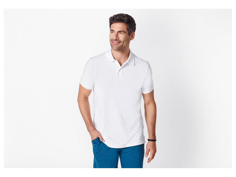 Mann in weißem Poloshirt und blauen Shorts, lächelnd, mit der Hand in der Tasche.