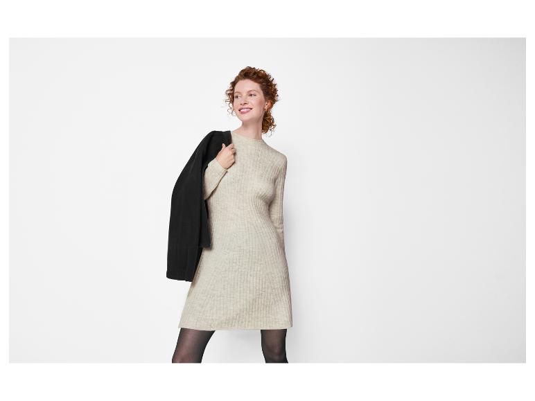 Rothaarige Frau in beigem Strickkleid mit schwarzer Jacke über der Schulter.