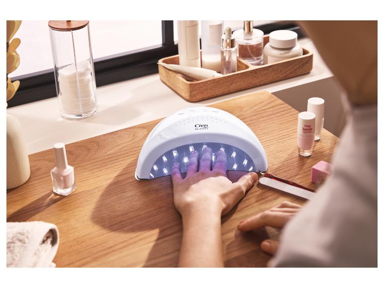 Frau trocknet Nägel in einer UV-Lampe, daneben Nagellacke und Maniküre-Zubehör.