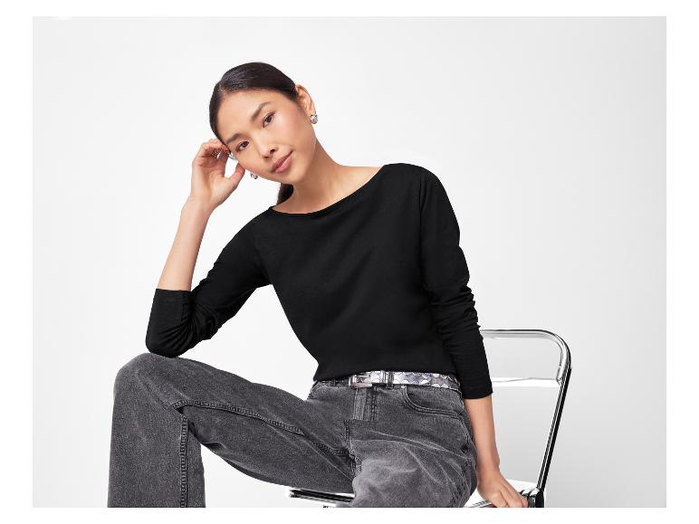 Frau in schwarzem Langarmshirt, grauen Jeans und silbernem Gürtel sitzt auf einem Stuhl.