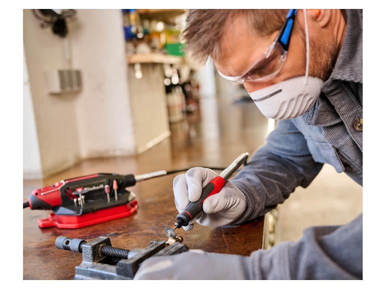 Mann in Schutzausrüstung verwendet ein Rotationswerkzeug für Detailarbeiten an einer Werkbank.
